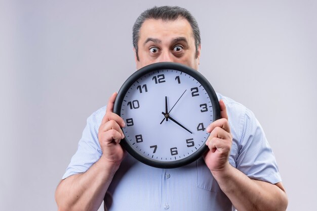 Shocked middle age man in blue striped shirt holding wall clock showing time while standing on a white background