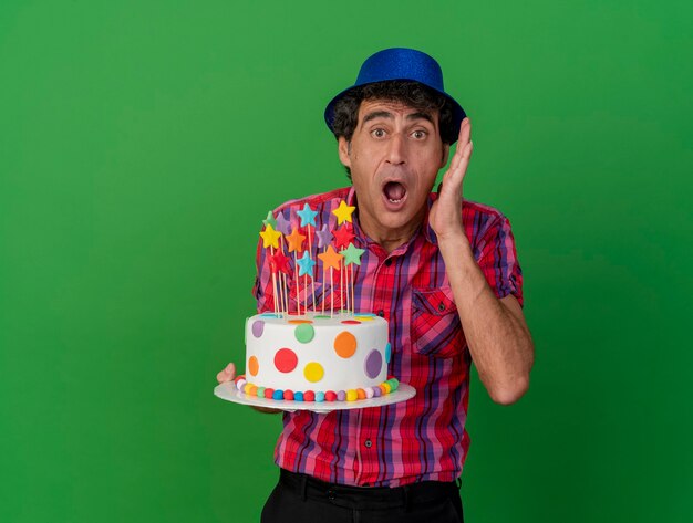 Шокированный кавказский тусовщик средних лет в шляпе с праздничным тортом, глядя в камеру, держа руку возле головы, глядя в камеру, изолированную на зеленом фоне с копией пространства