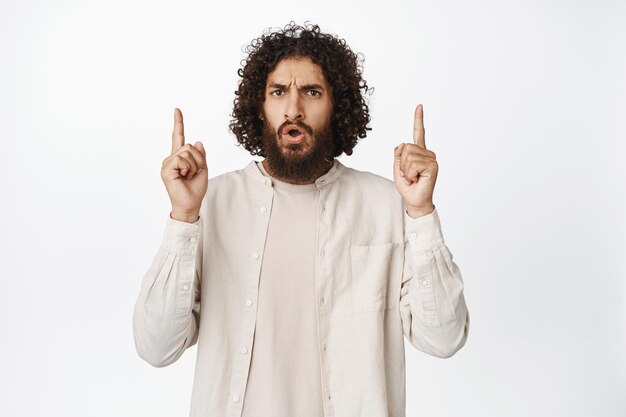 Shocked middle eastern guy pointing fingers up showing smth strange or freaky white background