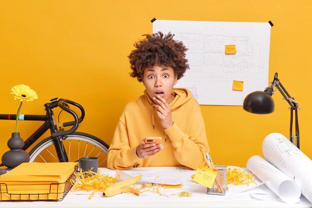 Shocked mixed race busy young woman stares surprised, uses smartphone, poses in coworking space has productive work at office desk surrounded by sketches and blueprints