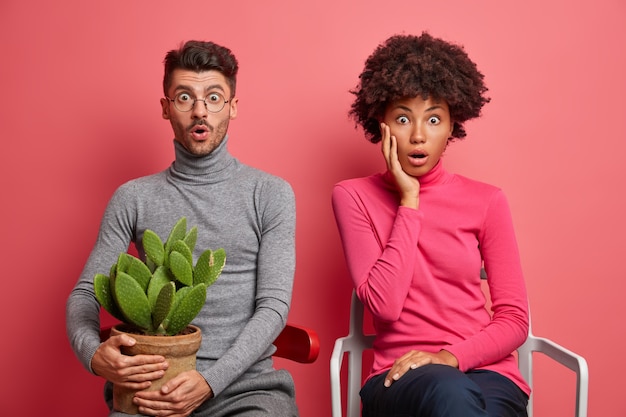 Shocked mixed race young woman and man sit next to each other impressed by shocking news pose on comfortable chairs dressed in casual clothing