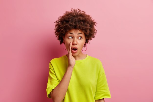 Shocked mysterious curly haired woman keeps hand near opened mouth tells secret spreads rumors whispers something secretly concentrated aside wears casual t shirt isolated over pink wall