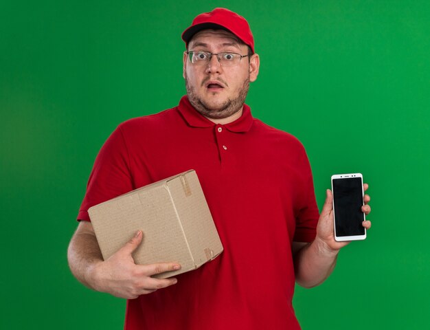 shocked overweight young deliveryman in optical glasses holding cardboard box and phone isolated on green wall with copy space