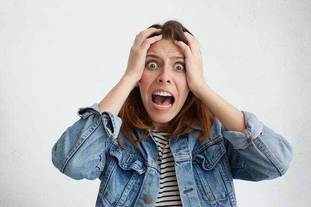 Free photo shocked panic caucasian girl dressed in stylish clothing holding hands on head and screaming in despair and frustration