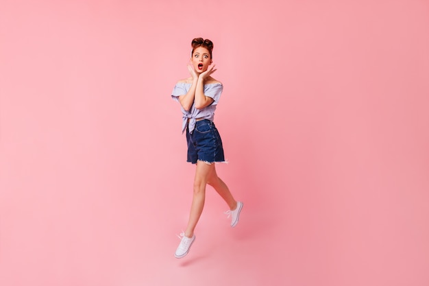 Shocked pinup girl in white shoes posing with open mouth. Full length view of emotional ginger woman jumping on pink space.