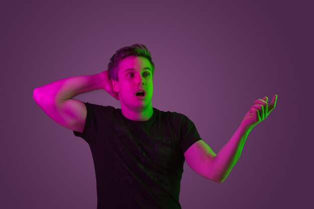 Shocked pointing at side. Caucasian man's portrait on purple studio background in neon light. Beautiful male model in black shirt. Concept of human emotions, facial expression, sales, ad. Copyspace.