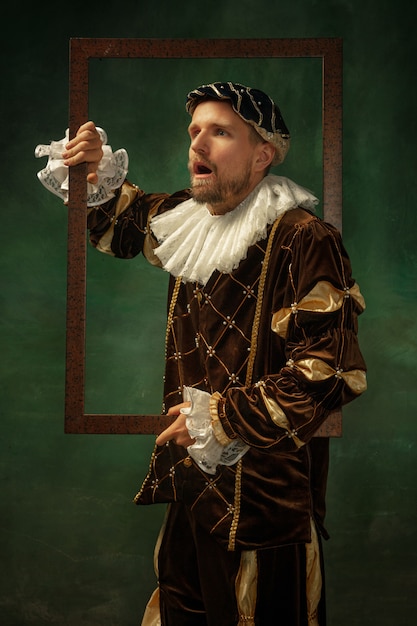 Shocked. Portrait of medieval young man in vintage clothing with wooden frame on dark background. Male model as a duke, prince, royal person. Concept of comparison of eras, modern, fashion, sales.