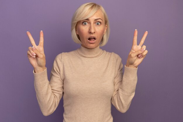 Шокированная симпатичная славянская блондинка жестикулирует знак рукой победы двумя руками на фиолетовом