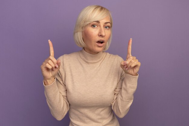 Shocked pretty blonde slavic woman pointing up with two hands on purple