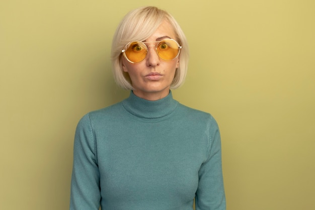 Shocked pretty blonde slavic woman in sun glasses looks at camera on olive green