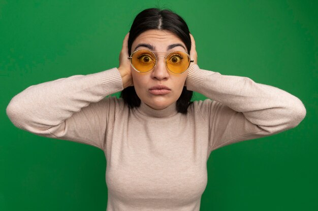 Шокированная красивая брюнетка кавказская девушка в солнцезащитных очках закрывает уши руками, глядя в камеру на зеленом