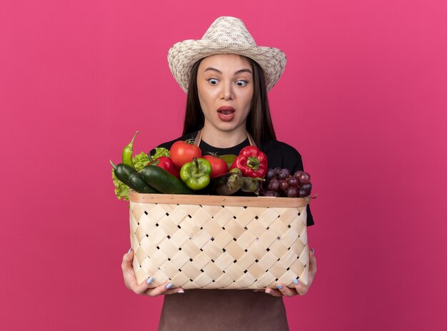 Shocked pretty caucasian female gardener wearing gardening hat holding and looking at vegetable basket isolated on pink wall with copy space