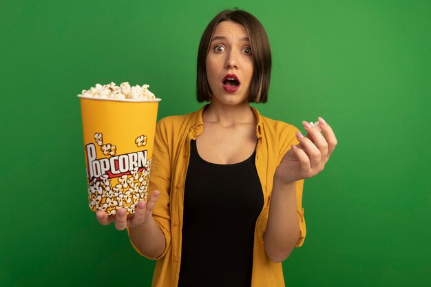 Shocked pretty caucasian woman holds bucket of popcorn on green
