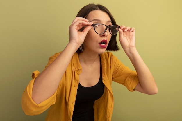 Шокированная красивая женщина держит и смотрит в сторону через оптические очки, изолированные на оливково-зеленой стене