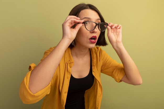 Shocked pretty woman holds and looks at side through optical glasses isolated on olive green wall