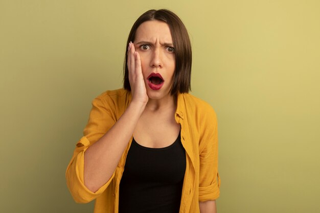 Shocked pretty woman puts hand on face isolated on olive green wall