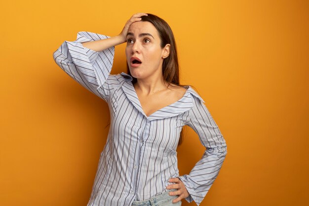 Shocked pretty woman puts hand on forehead and looks at side isolated on orange wall