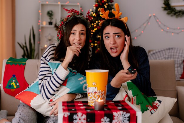 Una bella ragazza scioccata con la fascia da renna tiene il telecomando della tv seduto sulla poltrona con la sua amica che mangia popcorn godendosi il periodo natalizio a casa