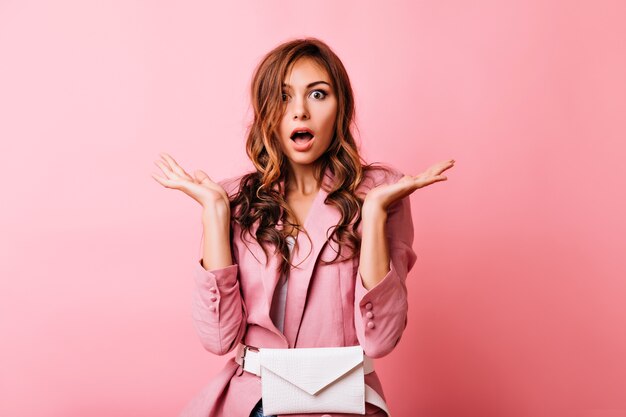 Shocked red-haired girl in pink jacket waving her hands. portrait of amazed fashionable woman isolated on pastel.