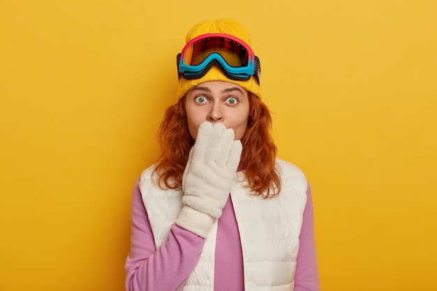 Shocked red haired woman covers mouth and stares at camera, wears special clothes and equipment for winter sport, enjoys skiing or snowboarding