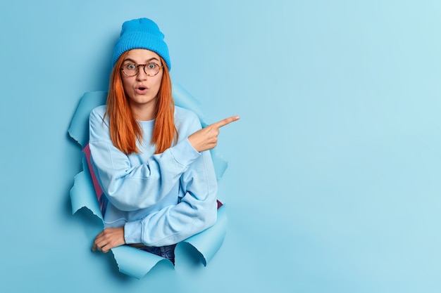 Shocked red haired young woman holds breath stares bugged eyes indicates away on copy space stands in ripped hole wears blue hat and jumper demonstrates something of high price on item