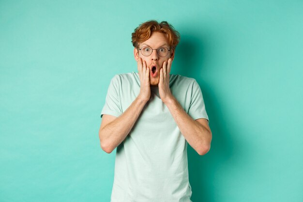 Shocked redhead guy in glasses checking out awesome promotion offer, gasping amazed and holding hands on face, staring at camera, mint background.