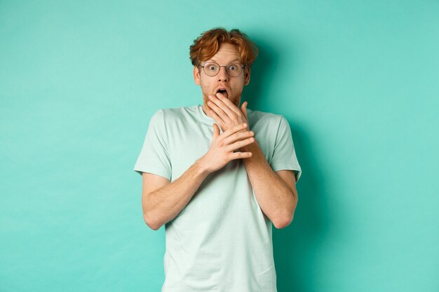 Shocked redhead man in glasses gasping startled, covering mouth and staring at camera scared, standing over turquoise background.