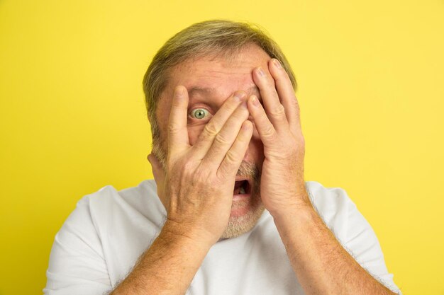 Shocked, scared, covering face with hands. Caucasian man portrait isolated on yellow studio background. Beautiful male model in white shirt.