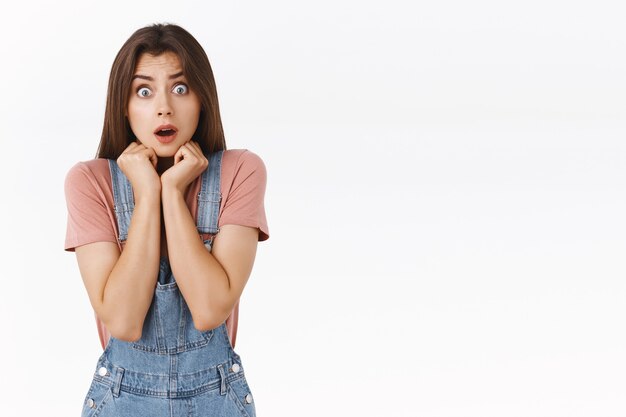 Shocked, scared timid insecure cute girl in dungarees and t-shirt, frightened staring camera feel unsafe and horrified from shocking news, shivering, press hands to chest terrified