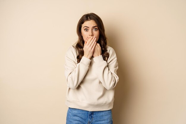 Shocked and scared woman cover mouth, looking stunned and speechless, worry about smth, standing over beige background