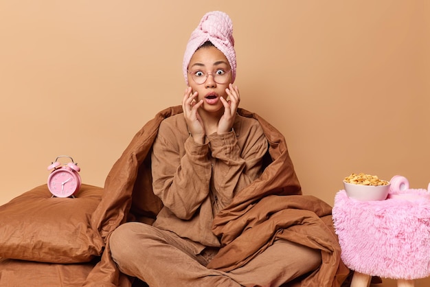 Free photo shocked scared woman in pajama and towel on head sits crossed legs on bed wrapped with blanket feels frightened after seeing nightmare going to have breakfast isolated over beige background