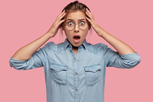 Free photo shocked scared woman remembers terrific moments, keeps both hands on head, keeps jaw dropped, wears denim jacket