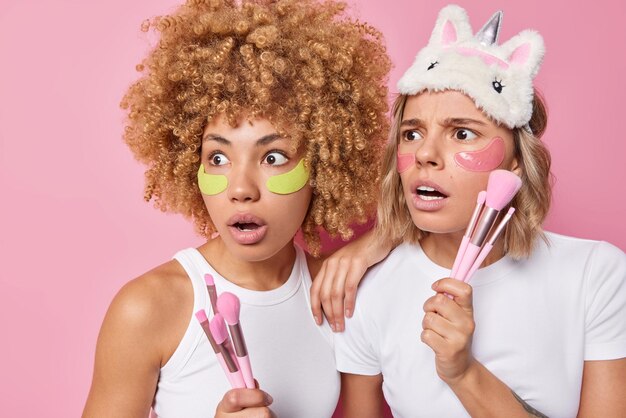 Shocked scared women stare away apply hydrogel patches under eyes pose with cosmetic brushes undergo beauty procedures dressed in casual white t shirts isolated over pink background Cosmetology