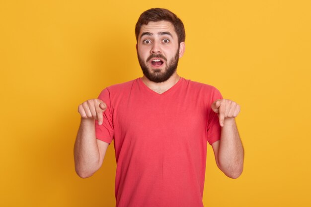 shocked scared young male being afraid of something, pointing down with his fore fingers, having surprised and astonished facial expression, posing isolated on yellow