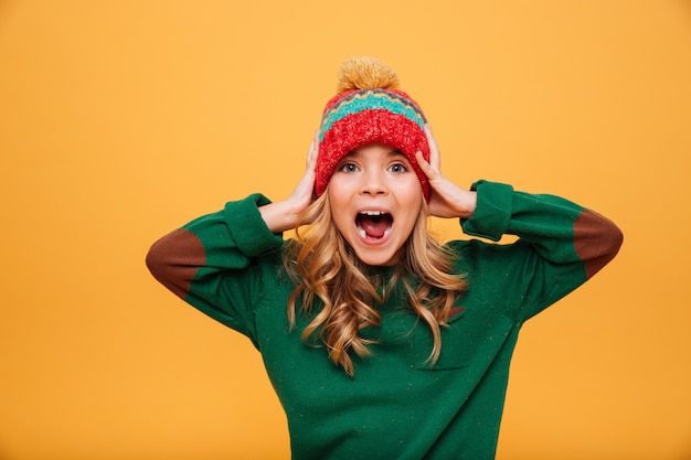 Shocked Screaming Young girl in sweater and hat holding her head while looking at the camera over orange