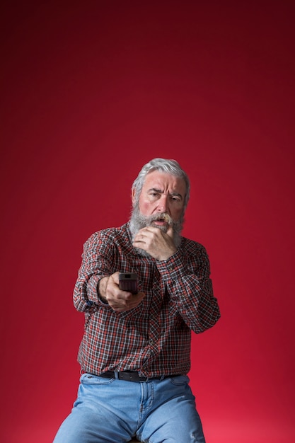 Shocked senior man changing the channel of television with remote control against red background