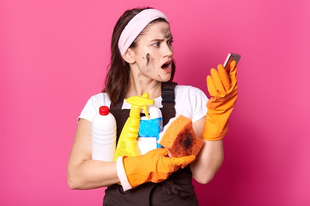 Потрясенная стройная брюнетка-домохозяйка держит бутылочки с моющим средством в одной руке и телефон в другой