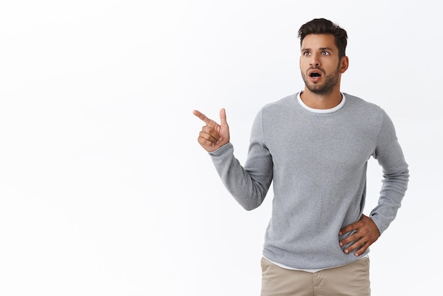 Shocked slightly worried handsome bearded caucasian guy in grey trendy sweater gasping drop jaw from amazement look concern pointing left shook and speechless check out something unusual