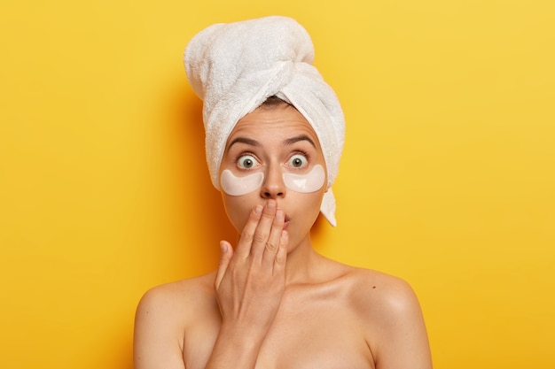 Shocked spa woman popping eyes, horrified by terrible relevation, nourishes undereye skin with beauty patches, wears wrapped towel on head, has anti wrinkle procedure, being naked.