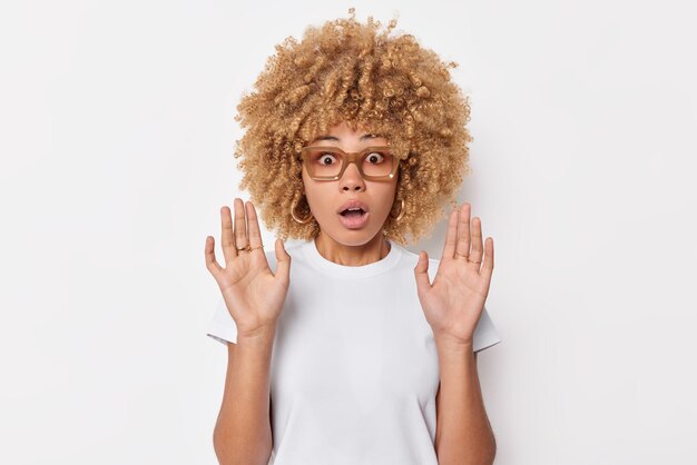 Shocked speechless curly haired woman keeps palms raised in defensive pose sees something terrifying holds breath from amazement wears spectacles and casual t shirt isolated over white background