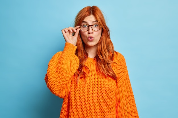 赤い髪のショックを受けた言葉のないヨーロッパの女性は、ニットのオレンジ色のジャンパーに身を包んだ大きな驚きで眼鏡を見つめ、予期しない感情から口を開きます。