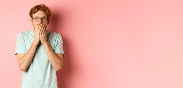 Shocked and speechless redhead guy covering mouth staring at camera startled standing in tshirt agai