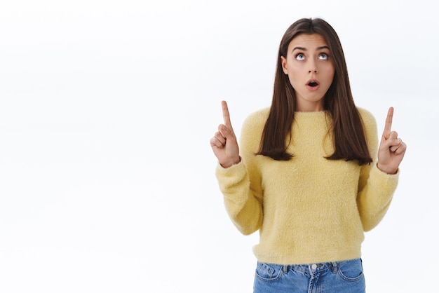 Free photo shocked speechless young brunette woman look and pointing fingers up with dropped jaw standing over white background in awe astounded with something huge or unbelievable copy space