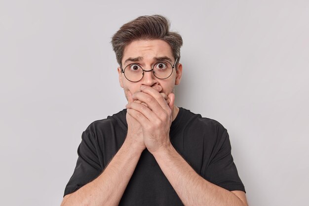 Shocked speechless young European man covers mouth with hands ties to be mute looks with fear at camera wears spectacles and casual t shirt poses against white background. Keep silence concept