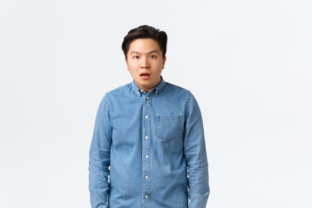 Shocked and startled asian male student, drop jaw and stare dumb at camera, cant understand what happening, feeling confused and puzzled, looking speehless over white background.