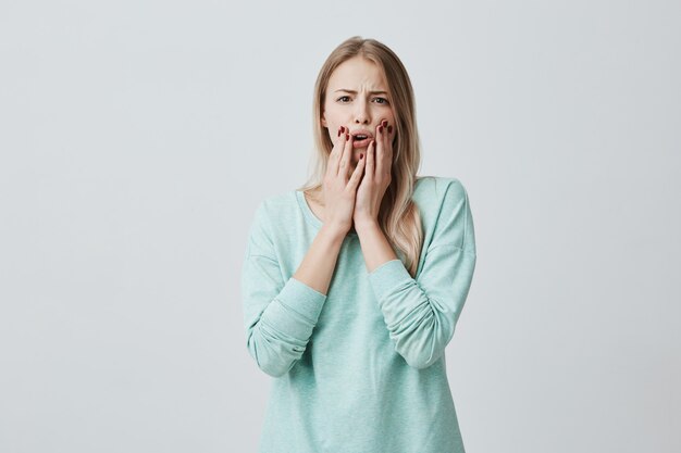 Shocked stunned blonde european woman with opened mouth, holding hands on cheeks, shocked by news she heard. Frightened terrified beautiful female model. Negative emotions