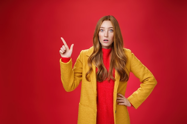 Shocked and stunned cute, confused ginger girl in yellow coat feeling worried and surprised open mouth lifting eyebrows amazed pointing at upper left corner, posing against red background.