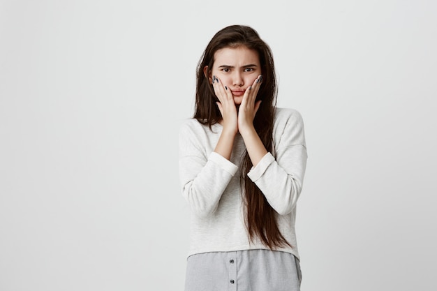 Shocked stunned emotional brunette teenage woman keeps hands on cheeks, being troubled