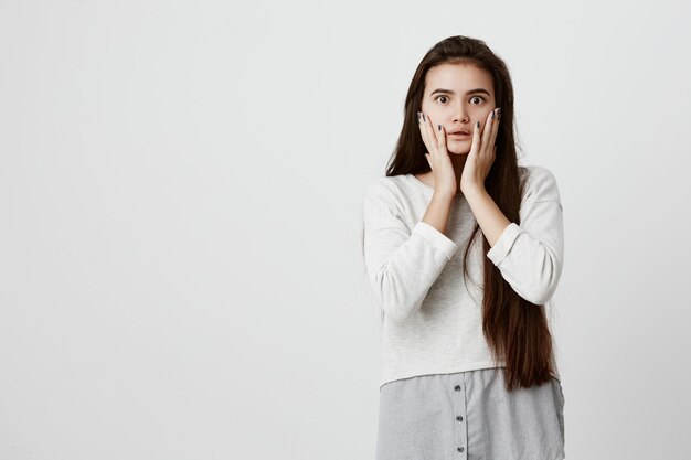 Shocked stunned emotional brunette woman keeps hands on cheeks, being troubled to listen advices of his parents. European young female with long dark hair can t believe in shocking news