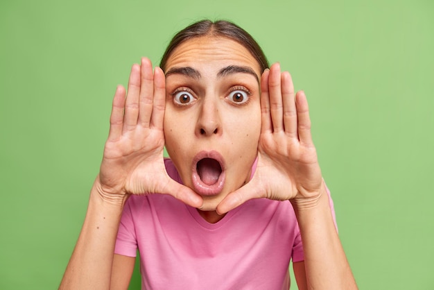 Free photo shocked stunned european woman keeps palms near face stares at camera with scared expression reacts on something very surprising keeps jaw dropped isolated over green wall